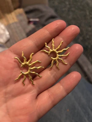 Solformade örhängen - örhängen i form av en sol med böljande strålar, tillverkade i stål  med guldtonad finish. Perfekt för dig som vill sticka ut med ett coolt och kreativt smycke. Säljes som par.
