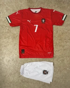 Portugal Ronaldo #7 fotbollströja Puma - Säljer en röd Portugal fotbollströja från Puma med nummer 7 och Ronaldo på ryggen. Tröjan har korta ärmar med gröna och vita detaljer vid mudden, samt ett subtilt mönster över hela tyget. Materialet är lätt och andas, perfekt för match eller träning. Ingår vita shorts med Portugal-emblem.