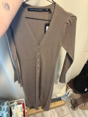 Ribbad långklänning  - Snygg ribbad långklänning i beige från 157 The Black Label Collection. Klänningen har v-ringning, långa ärmar och knappar hela vägen framtill. Perfekt för dig som gillar stilrena och bekväma plagg med en modern touch.