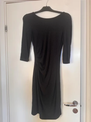 Svart/grå klänning från Vila - Supersnygg svart/grå klänning från Vila med drapering framtill som ger en smickrande midja. Modellen är knälång med trekvartsärm och har en diskret dragkedja i ryggen. 