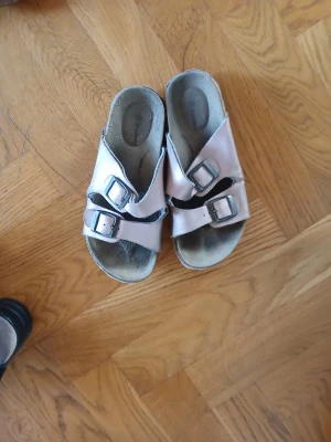 Rosa sandaler med spännen och glitter - Säljer ett par ljusrosa sandaler från Walkmaxx med två breda remmar och silvriga spännen. Sidan av sulan har en glittrig detalj som ger extra stil. Ovandelen är i syntet och sulan är korkliknande för skön komfort.