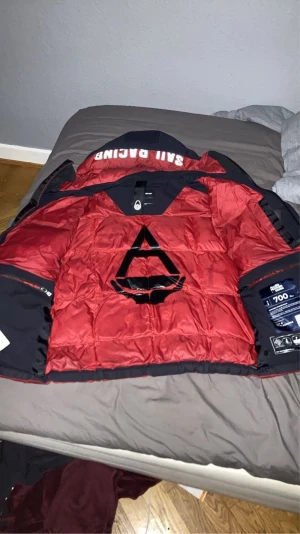 Sail Racing dunjacka röd och svart - Säljer en fet glacier jacka från Sail Racing i rött och svart med stor huva och coola loggor. Jackan har flera stora fickor, dragkedja och tryckknappar framtill.  Perfekt för kalla dagar.