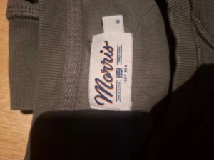 Olivgrön sweatshirt från Morris - Snygg olivgrön sweatshirt från Morris i storlek S. Tröjan har rund hals, långa ärmar och vit logotyp med brittisk flagga på bröstet. Tillverkad i mjuk bomull med ribbade muddar vid ärmslut och nederkant. Perfekt för chill dagar och streetwear.