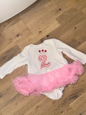 Vit och rosa tårtkjolsklänning 2 år Vit, rosa body med volang kjol - Supersöt vit klänning med långa ärmar och fluffig rosa tyllkjol. Framsidan har en stor rosa siffra 2 med prickar och en liten krona upptill, perfekt för födelsedagsfirande. Kjolen är volangig och ger en riktig prinsesskänsla.Vit body med lång ärm och en rosa tyllkjol. Bodyn har en applikation med siffran 2 och en krona. 🎉  använd endast 1 gång nypris: 29€