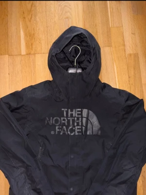 Svart vindjacka från The North Face - Svart vindjacka från The North Face med stor logga på bröstet. Jackan har huva, långa ärmar och två dragkedjeförsedda fickor framtill. Skick 8/10 ✅Skickar inom 48 timmar om inte samma dag 🚚
