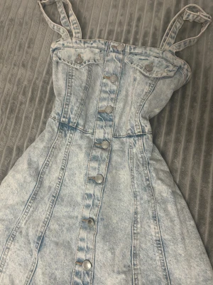 DENIM klänning  - Snygg ljusblå jeansklänning med justerbara axelband och knappar framtill. Klänningen har en figurnära passform och cool tvättad denim-look. Fickdetaljer upptill och dragkedja i sidan för enkel påklädning. Perfekt för dig som gillar retrovibbar och streetstyle.