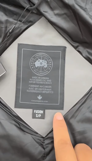 Grå pufferjacka från Canada Goose - Säljer en grå pufferjacka från Canada Goose med huva och snygg svart patch på ärmen. Jackan har två sidofickor, dragkedja och knappar framtill. Insidan är fodrad i svart och modellen är Fusion. Perfekt för kalla dagar och har en clean, modern look. ✅Kan lösa alla storlekar✅