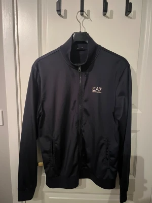 Svart trackjacka från EA7 Emporio Armani - Svart trackjacka från EA7 Emporio Armani med stilren design och logga på bröstet. Jackan har ståkrage, hel dragkedja framtill och två fickor med dragkedja. Materialet är mjukt och lätt polyester, perfekt för en sportig och clean look.