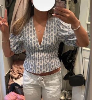Vit blus med blått mönster och puffärm - Säljer en vit blus med blått blommigt mönster och djup v-ringning. Blusen har puffiga korta ärmar och smockad midja som ger en snygg croppad passform. Perfekt för sommarens alla tillfällen, superfin till både jeans och kjol.
