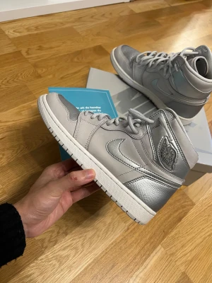 Nike Air Jordan 1 High Silver - Snygga Nike Air Jordan 1 High sneakers i silver och grått med metallic-detaljer. Skorna har klassisk snörning, rund tå och platt sula. Materialmix av skinn och syntet ger en lyxig vibe. Perfekt för dig som vill sticka ut med en ikonisk siluett.