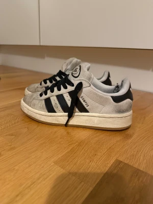 Adidas Campus grå sneakers mocka - Adidas Campus sneakers i grå mocka med svarta detaljer och klassiska tre ränder på sidan. Skorna har svart snörning, rund tå och vit sula. Adidas logga syns på tungan och hälen. Perfekt för dig som gillar retrostil och streetwear.