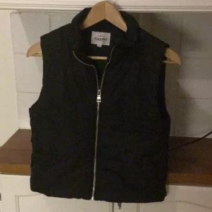 Svart dunväst från KappAhl - Svart dunväst från KappAhl med hög krage och silverfärgad dragkedja framtill. Västen har en enkel, stilren design och en reflexdetalj på ryggen. Perfekt för lager-på-lager under kyliga dagar.