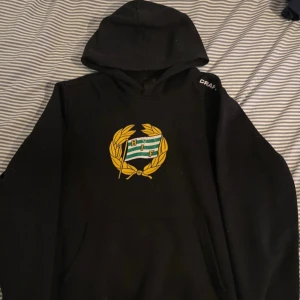 Svart Hammarby hoodie från Craft - Svart hoodie från Craft med Hammarby IF-emblem i grönt, vitt och gult på bröstet. Tröjan har huva, lång ärm och en stor magficka. Insidan är mjuk och fleecefodrad för extra komfort. Perfekt för dig som vill visa ditt stöd för Bajen.