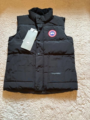 Svart CG väst - Svart dunväst med klassisk logga på bröstet. Västen har hög krage, två stora fickor framtill och knäppning med både dragkedja och tryckknappar. Perfekt för kalla dagar och har en stilren, quiltad design.Mycket skön och oanvändbar