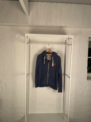 Blå hoodie från Superdry med dragkedja - Blå hoodie från Superdry Orange Label med vit snörning i huvan och röd logotyp på bröstet och ärmen. Tröjan har dragkedja framtill, två fickor och ribbade muddar. Materialet är mjuk bomull med fleece på insidan, perfekt för chilliga dagar.