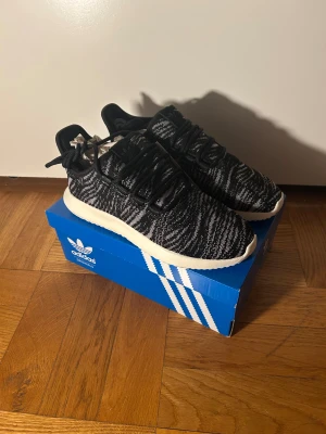 Adidas Tubular Shadow W sneakers - Adidas Tubular Shadow W sneakers i svart och vit med stickat mönster och rund tå. Snygg, låg modell med snörning och platt sula. Skon har en modern siluett och är tillverkad i syntetmaterial med textil för en cool streetstil. Strl 36.5, 38.5 och 40 finns i lagret.