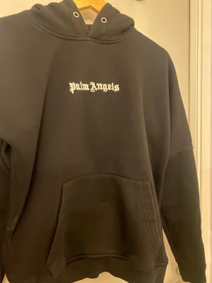 Svart Palm Angels hoodie med tryck - Svart hoodie från Palm Angels med stor vit logotyp på ryggen och mindre tryck framtill. Tröjan har huva med snörning, magficka och långa ärmar. Mjuk och skön, perfekt för streetwear-stilen. Passformen är loose och materialet känns som bomull.