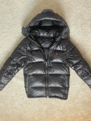 Svart dunjacka från Polo Ralph Lauren - Snygg svart dunjacka från Polo Ralph Lauren med glansig finish och röd logga på bröstet. Jackan har huva, dragkedja framtill och två sidofickor med dragkedja. Perfekt för kalla dagar. Köpt från zalando för 6500! Har kvitto 