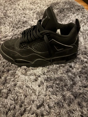 Svarta Air Jordan 4 Retro sneakers - Säljer ett par helsvarta Air Jordan 4 Retro sneakers med klassisk siluett och Jumpman-logga på plösen. Skorna har snörning, platt sula och är tillverkade i skinn och syntet. Grymma detaljer och ikonisk design, perfekt för dig som gillar streetwear.