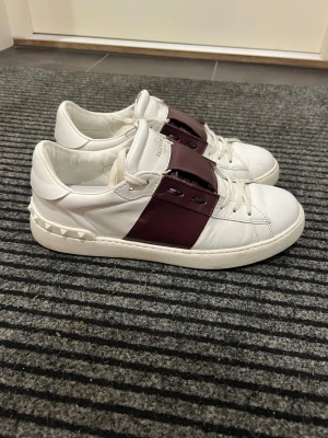 Valentino skor - Snygga Valentino Garavani Rockstud sneakers i vitt skinn med bred vinröd rem över snörningen och ikoniska nitar på hälen. Storlek 43, fits TTS. Använda ett dussintals gånger, inga funktionella defekter. Äldre modell med bastant kvalite, håller riktigt bra! BUD från 1499