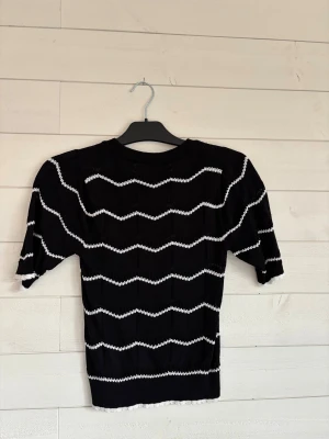 Svart stickad topp med vita ränder - Säljer en svart stickad topp från med vita vågiga ränder. Toppen har rund hals, korta ärmar och ribbad kant vid ärmslut och nederkant. Den är mjuk. Perfekt för dig som gillar att sticka ut lite extra.