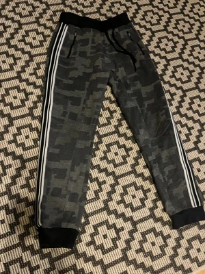 Gråa mjukisbyxor med snörning - Snygga mörkgrå mjukisbyxor med svart och grått camouflage-mönster, vita ränder längs sidorna och svarta muddar vid midja och benslut. Byxorna har snörning i midjan och dragkedjor vid fickorna. Använda men inte slitna