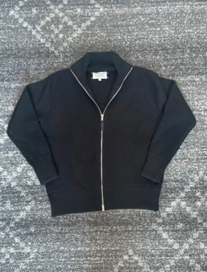 Svart kofta från Maison Margiela - Svart ribbstickad kofta från Maison Margiela med hel dragkedja framtill och hög krage. Tröjan har långärmad design och klassiska vita stygn i nacken. Perfekt för dig som gillar stilrena och minimalistiska plagg med exklusiv känsla.