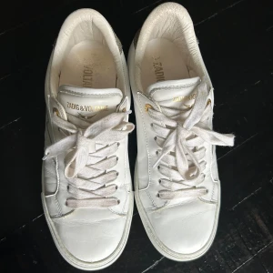 Vita skinn sneakers från Zadig & Voltaire strl.39 - Snygga vita sneakers från Zadig & Voltaire i skinn med cool blixtdetalj på sidan och glittrig detalj på hälen. Skorna har snörning med vita skosnören och guldfärgade detaljer vid snörhålen. Perfekta för dig som vill ha en clean men ändå unik look.strl.39