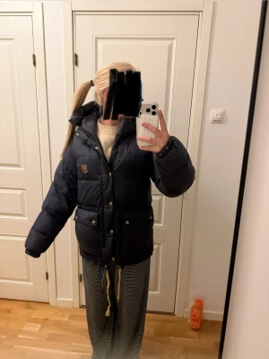 Mörkblå pufferjacka från Fjällräven XS - Säljer en mörkblå pufferjacka från Fjällräven i storlek XS. Jackan har huva, guldfärgade knappar, dubbla dragkedjor och flera fickor. Materialet är syntet och jackan är quiltad för extra värme. Perfekt för kalla dagar och har en klassisk Fjällräven-logga på bröstet. Den har lite sminkfläckar i luvan som jag dock aldrig har försökt få bort. Även ett litet hål vid dragkedjan som dock är på insidan av jackan.