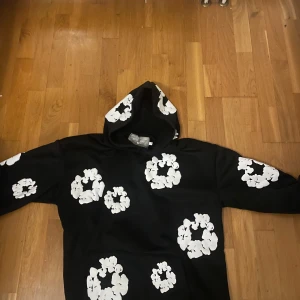 Svart hoodie från Denim Tears med blommor - Svart hoodie från Denim Tears med stora vita blommönster över hela tröjan. Tröjan har huva, lång ärm och en klassisk känguruficka framtill. Perfekt för dig som gillar streetwear och vill sticka ut med en unik design.