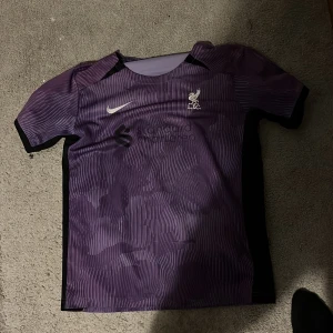 Lila Liverpool Nike fotbollströja - Liverpool FC fotbollströja från Nike i lila med vita detaljer och klubbmärke på bröstet. Tröjan har ett randigt mönster och korta ärmar. Materialet är lätt och andas, perfekt för träning eller match. Standard Chartered-logga på framsidan.