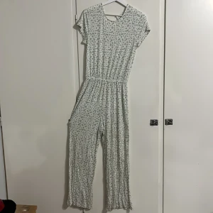 Blommig jumpsuit från Kappahl - Supersöt jumpsuit från Kappahl i storlek 164. Ljusgrön och vit med blommigt mönster, korta ärmar och v-ringad rygg. Mjuk och skön i bomull, med resår i midjan för en snygg passform. Perfekt för dig som gillar avslappnad och trendig stil.