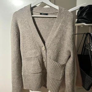 Beige stickad kofta från Gina Tricot - Mysig beige stickad kofta från Gina Tricot med v-ringning och tre knappar framtill. Koftan har två framfickor och ribbade muddar vid ärmslut och nederkant. Perfekt för lager-på-lager och chill dagar.