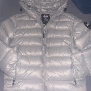 Silver dunjacka från Canada Goose S - Säljer en riktigt snygg silverfärgad dunjacka från Canada Goose. Jackan har en glansig finish, huva, dragkedja framtill och två sidofickor med dragkedja. Klassisk Canada Goose-logga på ärmen och reflekterande detalj på huvan. Perfekt för kalla dagar.