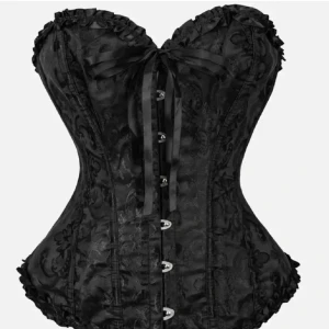 Svart korsett med snörning och rosett - Snygg svart korsett med vackert mönstrad yta, snörning framtill och dekorativ rosett vid bysten. Korsetten har markerad midja och knäppning framtill, samt volangkant upptill och nedtill för extra detalj.