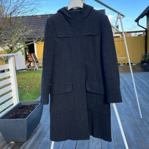 Mörkgrå kappa med huva från Lindex - Snygg mörkgrå kappa från Lindex i storlek 40. Modellen har huva, stora fickor framtill och stängs med både dragkedja och knappar. Tillverkad i ullblandning, perfekt för kyliga dagar. Stilren och enkel design som funkar till många olika looks.