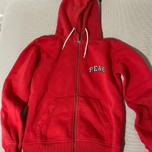 Röd hoodie från Peak Performance - Röd hoodie från Peak Performance med dragkedja, huva med vita snören och broderad PEAK-logga på bröstet. Mjuk bomullskänsla, kängurufickor och ribbade muddar.  