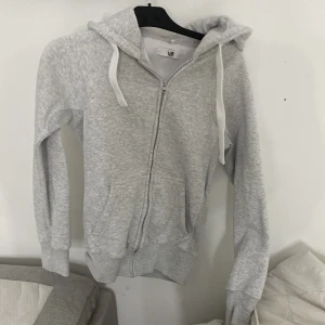 Ljusgrå hoodie med dragkedja XS - Mysig ljusgrå hoodie från Russemerch i storlek XS. Tröjan har huva med vita snören, hel dragkedja framtill och två fickor. Tillverkad i mjuk bomullsblandning med fleecefoder på insidan. Perfekt för kyliga dagar. Säljer då den inte kommer till användning, använd ca 3 ggr😊