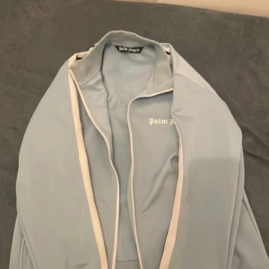 Ljusblå vindjacka från Palm Angels - Snygg ljusblå vindjacka från Palm Angels med vit piping längs dragkedjan och logga på bröstet. Jackan har en sportig look och är perfekt för dig som gillar streetwear. Lätt material och klassisk passform.
