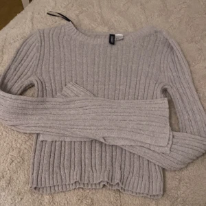 Ljusgrå ribbstickad tröja h&m - Supermjuk ljusgrå ribbstickad tröja från h&m med lång ärm och rund hals. Tröjan har en snygg, lite croppad passform och breda ribbade detaljer som ger en trendig look. Perfekt att styla med jeans eller kjol för en chill vibe. Aldrig använd helt ny 