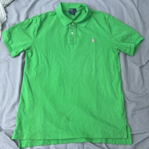 Grön pikétröja från Ralph Lauren - Snygg grön pikétröja från Polo Ralph Lauren med klassisk krage och två knappar framtill. Tröjan har korta ärmar och en rosa broderad logga på bröstet. Tillverkad i mjuk bomull som är skön mot huden. Perfekt för dig som gillar färg och stil.