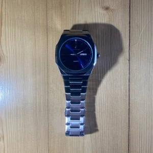 Blå kvartsarmbandsur från  - Snygg armbandsklocka från  med en djupblå urtavla och dag/datumvisning. Klockan har en robust, kantig boett och länkarmband i silverfärgat metall. Perfekt för dig som gillar stilrena accessoarer med lite edge.