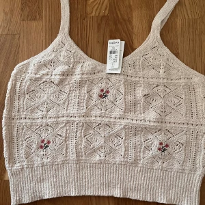 Beige stickad linnetopp från Kappahl - Säljer en beige stickad linnetopp från Kappahl i storlek XL. Toppen har ett snyggt hålmönster och broderade blommor i rött och grönt framtill. Den är ärmlös med smala axelband och har en ribbad kant nedtill. Perfekt för sommarens alla dagar!