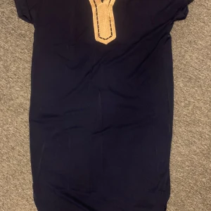 Mörkblå klänning med guldiga detaljer - Mörkblå klänning i rak modell med korta ärmar och snygga guldiga virkade detaljer längs halsringningen. Enkel och stilren design som ger en elegant touch. Perfekt för dig som gillar klassiska färger med en twist.