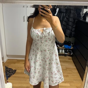 Vit blommig kortklänning med snörning - Superfin vit klänning med rosa och gröna blommor, smala axelband och söt snörning framtill. Klänningen har en lätt utställd form och är perfekt för varma dagar. Den är kort och har en romantisk vibe med sitt blommiga mönster och tunna material.