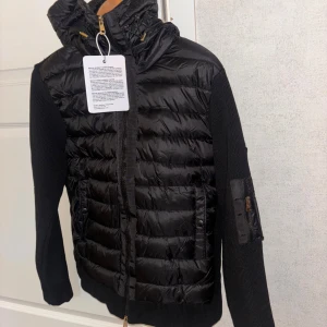 Svart Moncler pufferjacka med huva - Svart pufferjacka från Moncler med stickade ärmar och quiltad front. Jackan har huva med snörning, dragkedja framtill och fickor med tryckknappar. På ena ärmen finns en ficka med dragkedja och Moncler-logga. Perfekt för kyliga dagar.