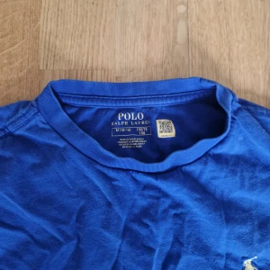 2 st Blå t-shirt från Polo Ralph Lauren - Snygg blå t-shirt från Polo Ralph Lauren med klassisk rund hals och korta ärmar. Ikoniska broderade loggan i vitt på bröstet. Tillverkad i mjuk bomull som är skön mot huden. Perfekt för en clean och stilren look.