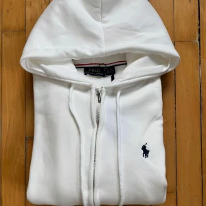 Vit hoodie från Polo Ralph Lauren - Vit hoodie från Polo Ralph Lauren med dragkedja och huva. Tröjan har långa ärmar, snörning vid huvan och klassisk Polo-logga broderad i marinblått på bröstet. Tillverkad i mjuk bomull för skön komfort. Perfekt för chill dagar.