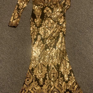 Svart långklänning med guldpaljetter - Superelegant svart långklänning med glittrande guldpaljetter i ett detaljerat mönster. Klänningen har långa ärmar, figurnära passform och dragkedja i ryggen. Perfekt för dig som vill sticka ut med en lyxig look. Använd endast en gång!