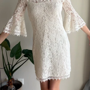 Vit spetsklänning från Colcci (Ibiza brand)  - Superfin vit kortklänning från Colcci (Ibiza brand) i spets med blommigt mönster. Offshoulder-modell med vida trekvartsärmar och dekorativ kant upptill. Perfekt för dig som vill ha en romantisk och trendig look. 